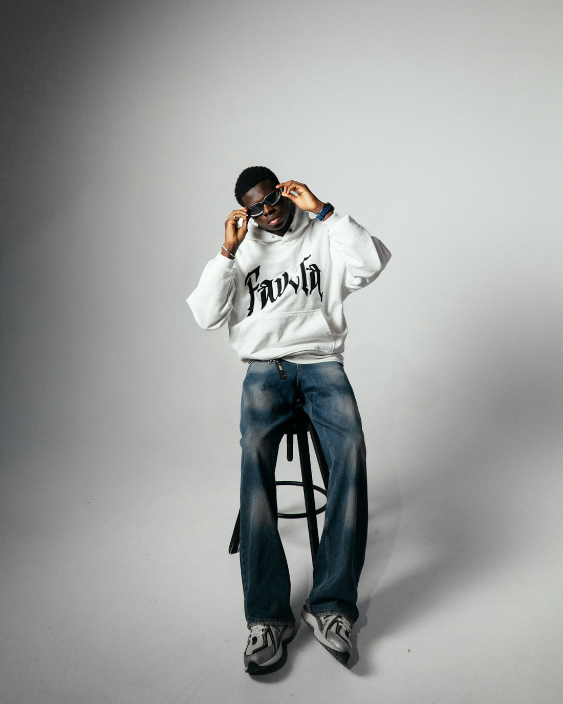 Man in hoodie and jeans adjusts sunglasses while sitting.